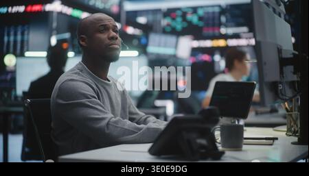 Male Stock Broker Analyzing Complex Trading Data at Multi Monitor Desk. Focused Black Man Scrutinizing Fluctuating Financial Graphs and Indices on Displays. Expert Workforce in Brokerage Firm. Stock Photo