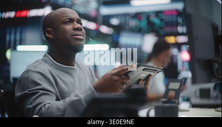 African American Investor Tracking Asset Performance in Innovative Trading Center. Man Focused on Digital Dashboard on Monitors and Digital Tablet. Intense Concentration in Global Finance Industry. Stock Photo