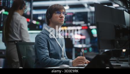 Young Male Financial Analyst in Glasses and Headset Sits at Modern Workstation, Gazes at Large Trading Monitors, Tracking Real Time Price Movements During Critical Market Session. Digital Monitoring. Stock Photo