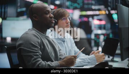 Seasoned African American Financial Expert Provides Hands on Training to Young Male Colleague. Men Deeply Engaged in Analyzing Real Time Data on Digital Tablet and Monitors. Mentorship, Tech Training. Stock Photo