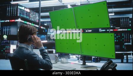 Young Man Speaks on Smartphone at Multi Monitor Trading Desk With Green Screen Displays, Analyzing Market Data, Coordinating Strategy, Managing Global Finance Operations in Digital Exchange Hub. Stock Photo
