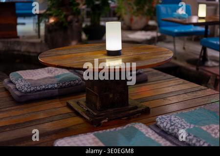 Wooden low table with modern lamp surrounded by floor cushions for seating at cozy outdoor lounge at night. Warm light creates relaxing atmosphere ide Stock Photo