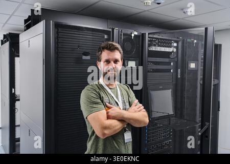 IT technician stands with arms crossed in a secure data center server room surrounded by server racks and networking equipment, conveying confidence, professionalism and modern technology infrastructure. || Model released Stock Photo