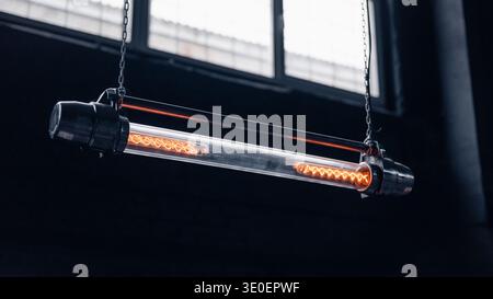 Suspended filament tube lamp. Industrial tubular pendant light suspended by chains, showcasing two glowing filaments inside a clear glass housing. Stock Photo