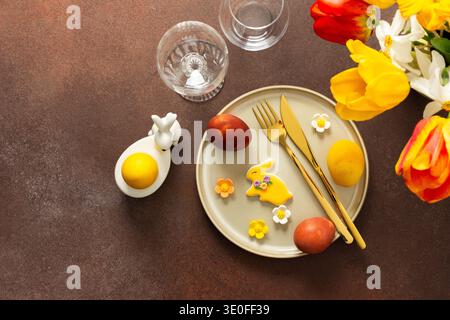 A beautifully arranged Easter table displays vibrant eggs, elegant gold cutlery, and a lively bouquet of tulips and lilies. This setting creates a cheerful atmosphere for the holiday feast Stock Photo