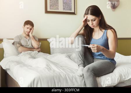 Upset woman sitting on bed and looking at pregnancy test Stock Photo