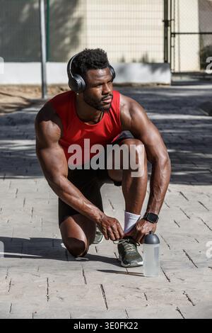 In a lively public park, an African-American man kneels to tie his shoelaces while getting ready for his sports activity. He wears athletic gear and h Stock Photo