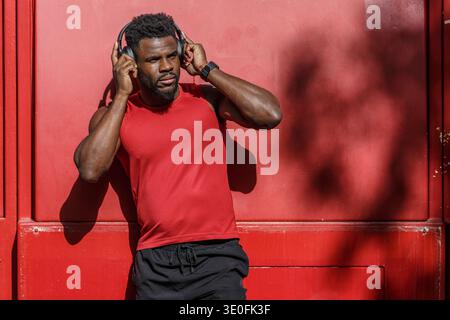 A man leans against a vibrant red wall, wearing headphones after completing an intense workout session. He appears relaxed and immersed in his music, Stock Photo