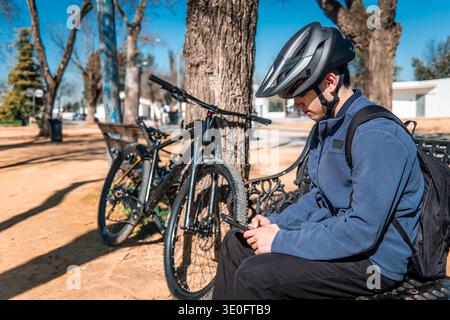 Young man wearing a helmet and backpack sitting on a bench, checking his cellphone next to his bicycle in a park Stock Photo