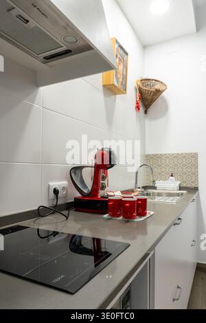 Contemporary kitchen featuring a coffee maker, induction hob, sink, and red mugs on a gray countertop with white wall tiles Stock Photo