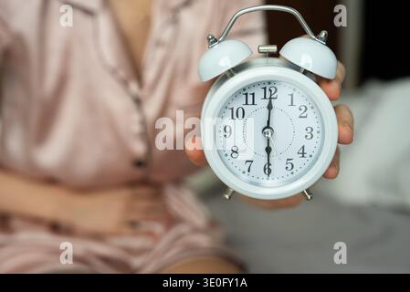 A woman in pink pajamas holds a white alarm clock in her hand, sitting in bed, illustrating a morning routine or time management concept with soft foc Stock Photo