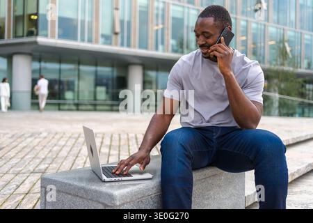 African American man freelancer using laptop talking on phone on urban street in city. Business man having virtual meeting. Professional programmer using phone laptop working outdoors. Remote work Stock Photo