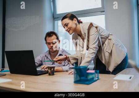 Business colleagues collaborate at a desk, woman leaning over to discuss work with man on laptop Stock Photo