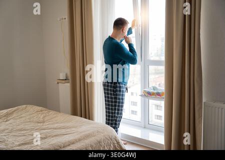 A young caucasian 35 old caucasian man looks out the window after waking up and collects his thoughts in apartment. Lifestyle and planning Stock Photo