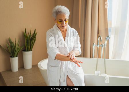 Mature adult old woman applying moisturizing body lotion on hands. Senior lady rubbing nourishing anti age cream doing morning daily skincare routine in bathroom. Beauty treatment spa body skin care Stock Photo