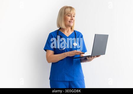 Senior female healthcare professional wearing blue scrubs and stethoscope working on laptop Stock Photo