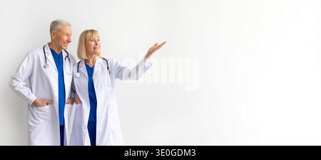 Two mature doctors in white coats standing on white background, pointing aside Stock Photo