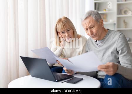 Concerned senior couple reading financial documents at home while sitting with laptop Stock Photo