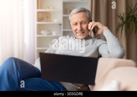 Smiling senior man relaxing on sofa, talking on smartphone and using laptop Stock Photo