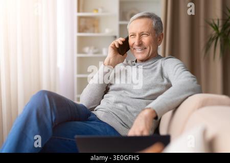 Happy senior man sitting comfortably on sofa at home talking on smartphone Stock Photo