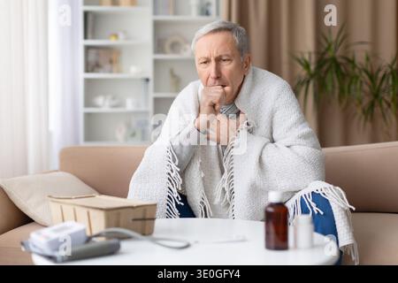 Senior man sitting on sofa wrapped in blanket while coughing at home Stock Photo