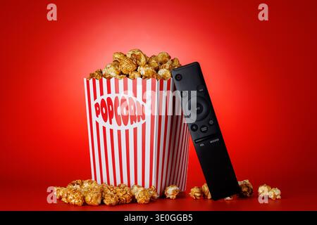 Popcorn in paper bucket with TV remote on bright red background, movie night entertainment concept Stock Photo