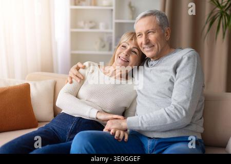 Senior couple sitting closely together on sofa at home looking away thoughtfully Stock Photo