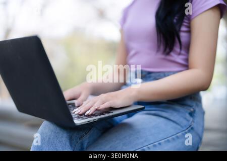 Adult Asian woman types laptop computer outdoors. Focus work study connect internet anywhere. Modern tech remote business learning. Casual productive Stock Photo