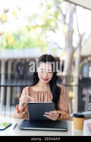 Focused young Asian woman using digital tablet and stylus at outdoor cafe Remote work student creator enjoying technology productivity Stock Photo