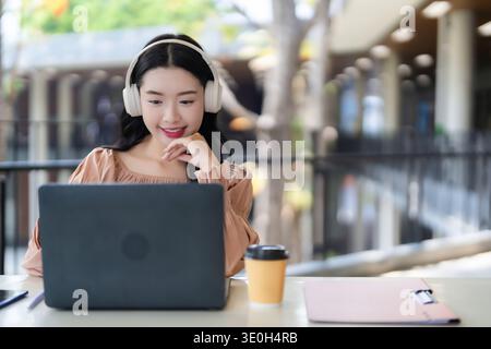 Asian adult woman engaged remote work or study using laptop headphones outdoor cafe enjoying technology and modern lifestyle Stock Photo
