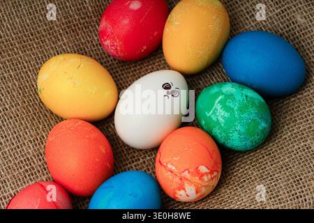 Sad white egg with hand-drawn face among colorful Easter eggs on burlap.  Concept representing being different, social exclusion, sadness during holid Stock Photo