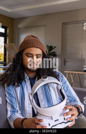 Man sitting on gray sofa wearing brown beanie and holding white VR headset near window light. Minimalist, immersive, technology, home, lounge, curious Stock Photo