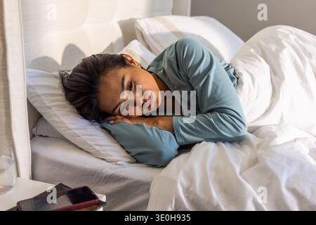 Asian woman lying on right side on bed wearing light blue loungewear, bedside phone and water. Bedroom, restful, serene, cozy, natural, soft, pillows Stock Photo