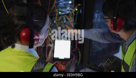 Diverse coworkers wearing yellow vests and red earmuffs, testing cables with tablet in data center. Serverroom, rack, technician, monitor, diagnostics Stock Photo
