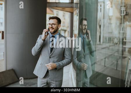 A businessman is standing outside a modern building. He is wearing a suit and glasses. He is talking on his phone with a slight smile. The glass refle Stock Photo