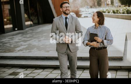 Two colleagues are walking together on a city sidewalk. They are discussing work and enjoying the sunny afternoon. The man is wearing a suit while the Stock Photo