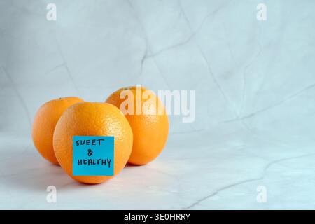 Fresh whole oranges arranged on a light background with a blue sticky note featuring the handwritten text sweet and healthy Stock Photo