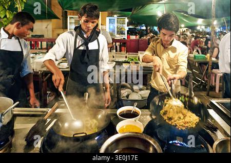 Chefs cook street food in woks as steam rises in Siem Reap night market. The busy scene shows hands working to serve customers. Stock Photo