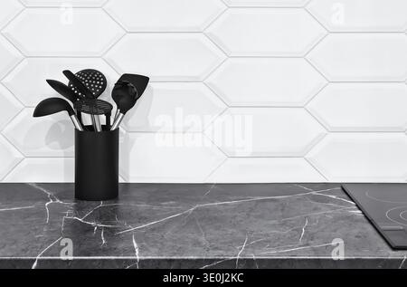 Modern minimalist kitchen interior featuring a sleek white marble countertop and a geometric-patterned white wall. A container of black dishware provi Stock Photo
