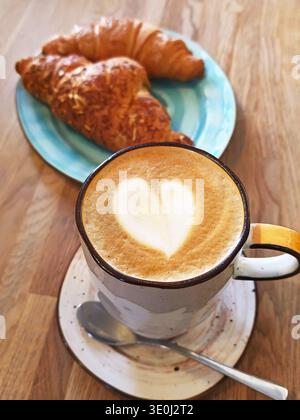 Cup of latte with heart-shaped foam art served with fresh croissant on a rustic wooden table. Cozy breakfast setting with warm morning atmosphere and Stock Photo