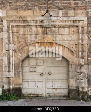 Weathered stone archway with an old, distressed white wooden door and metal ring handles. Historic architecture featuring rough textures, an ancient feel, and green plants at the base Stock Photo