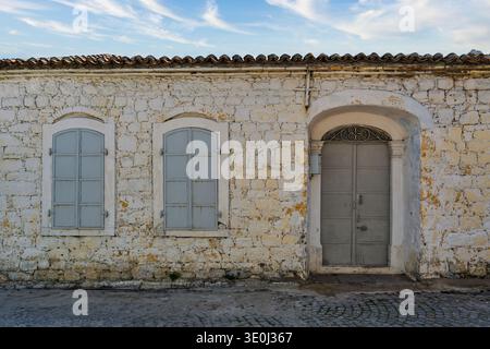 The scene shows a worn stone wall with a gray door and two gray windows. The sun highlights the textures of the wall. The sky shows some clouds, adding to the daytime atmosphere Stock Photo