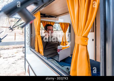 Man talking on smartphone, working remotely on laptop inside a camper van, enjoying nomadic lifestyle and travel Stock Photo