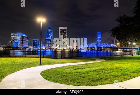 Vibrant night view of downtown Jacksonville, Florida, showcasing the illuminated city skyline, Main Street Bridge, and reflections on the Saint Johns River with a scenic park path Stock Photo