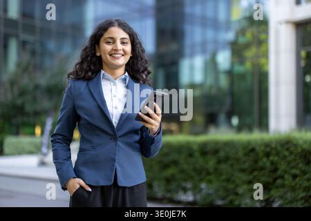 Young businesswoman with dark curly hair smiles confidently, holding a smartphone with one hand in pocket before a modern glass office building, symbolizing ambition and digital connectivity Stock Photo