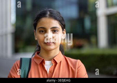 Young indian woman student smiling at camera, wearing orange shirt and backpack, standing confidently on a city campus outdoors head and shoulders portrait with blurred background Stock Photo