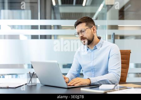 Business man wearing glasses and a light blue shirt concentrating on his laptop keyboard, working at a contemporary desk in a bright office environment Stock Photo