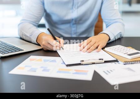 Businessman using a pen to sign a form on a clipboard, reviewing financial documents, graphs, and a calculator on a desk, representing accounting, analysis, and data management Stock Photo