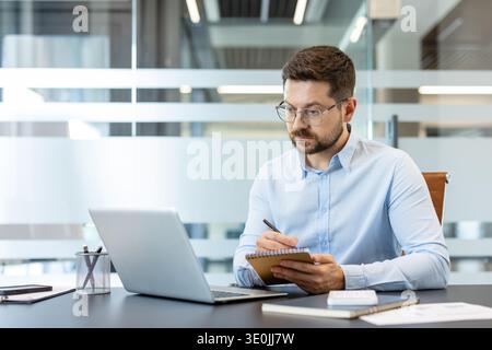 Businessman in glasses and a light blue shirt actively taking notes in a spiral notebook while focusing on his laptop computer, working diligently at a modern office desk Stock Photo