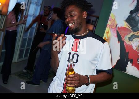 Young Black Man Smiling While Using Smartphone and Holding Beer Bottle Stock Photo